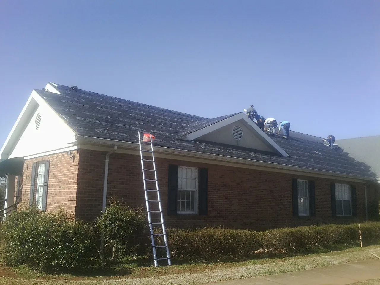 Crew tearing shingles off a brick ranch roof with a ladder set — Metal Roof Repair work by our crew in Humble