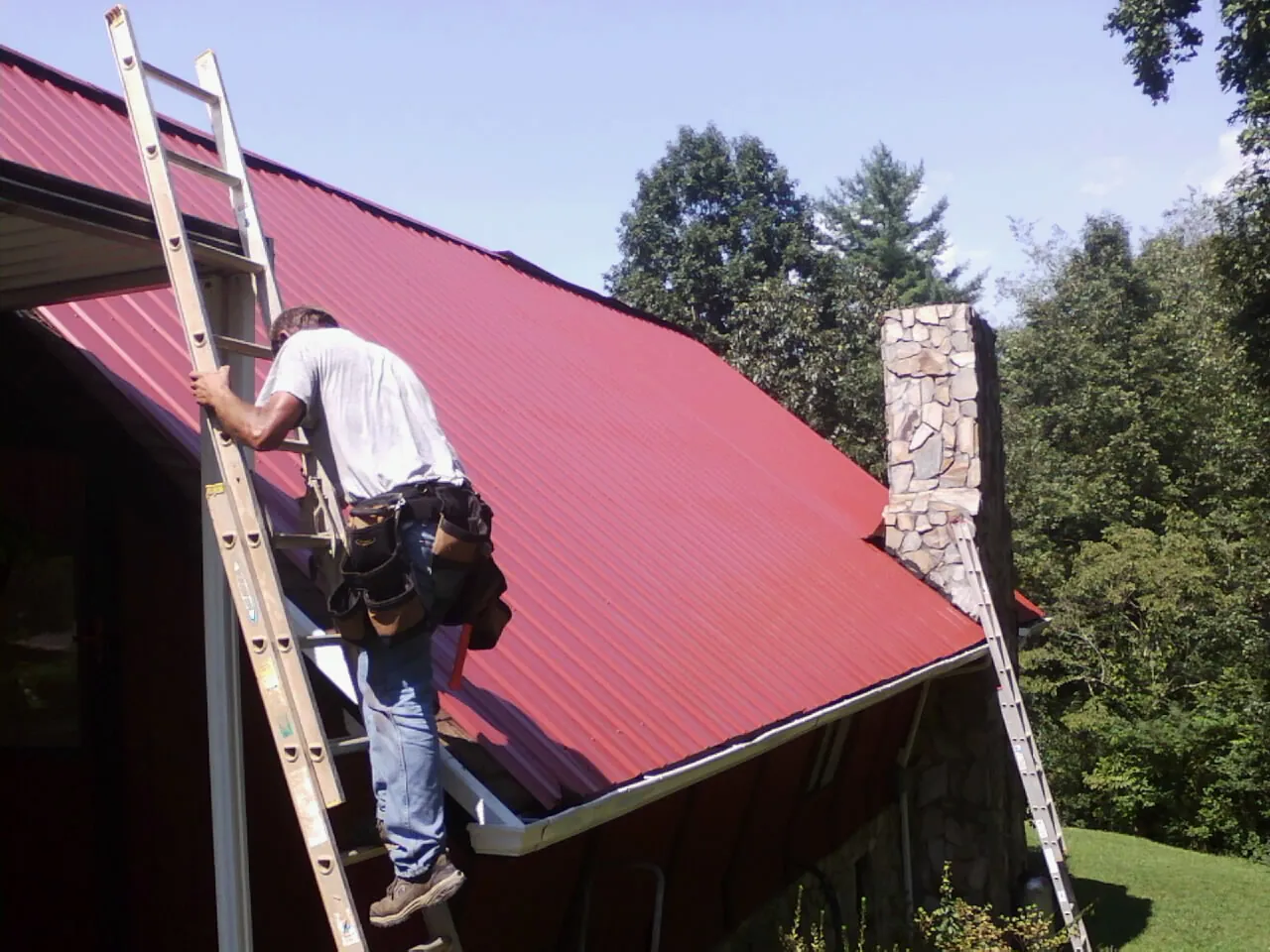 Roofer on a ladder beside a red metal roof and stone chimney — Soffit & Fascia Repair by Roofing Brothers in Humble